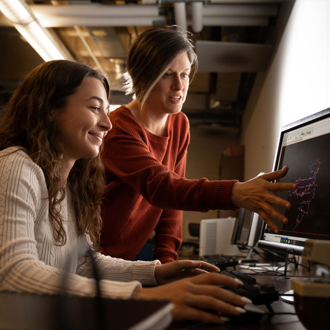 A Female professor stands next to a seated student at a computer and holds her hand up to the computer monitor to point something out for the student.