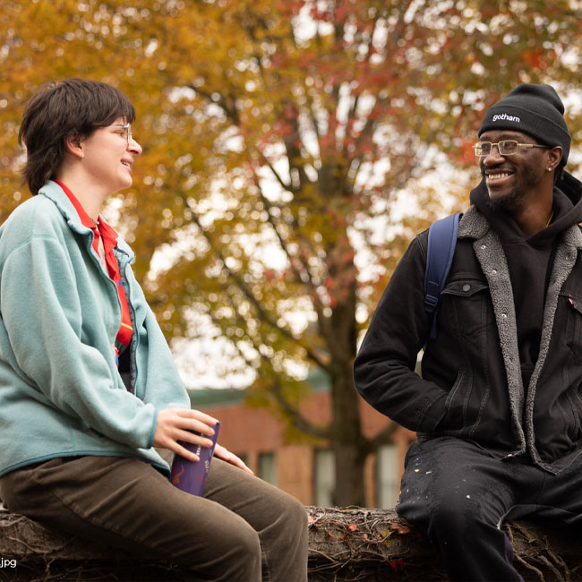 2 students sit on a rock wall under a tree with colored leaves and enjoy a good conversation outside.