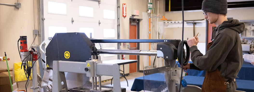 A young male wearing a safety apron works on a manufacturing machine in a warehouse setting with a garage door in front of him.