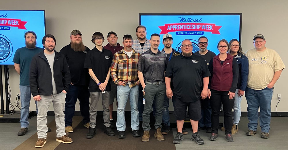Apprentices at Amphenol Aerospace pose for a picture in front of large tv monitors that show National Apprentice Week logos on them.
