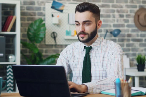 Male in shirt and tie works at a laptop in creative media office.