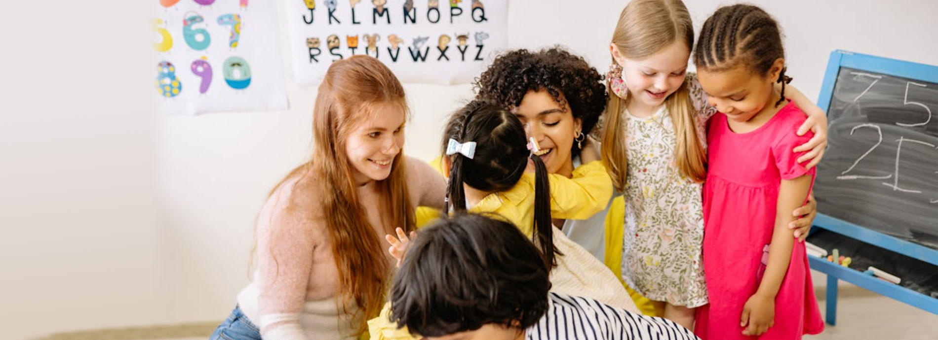 Female teacher sits with young kids on classroom carpet.