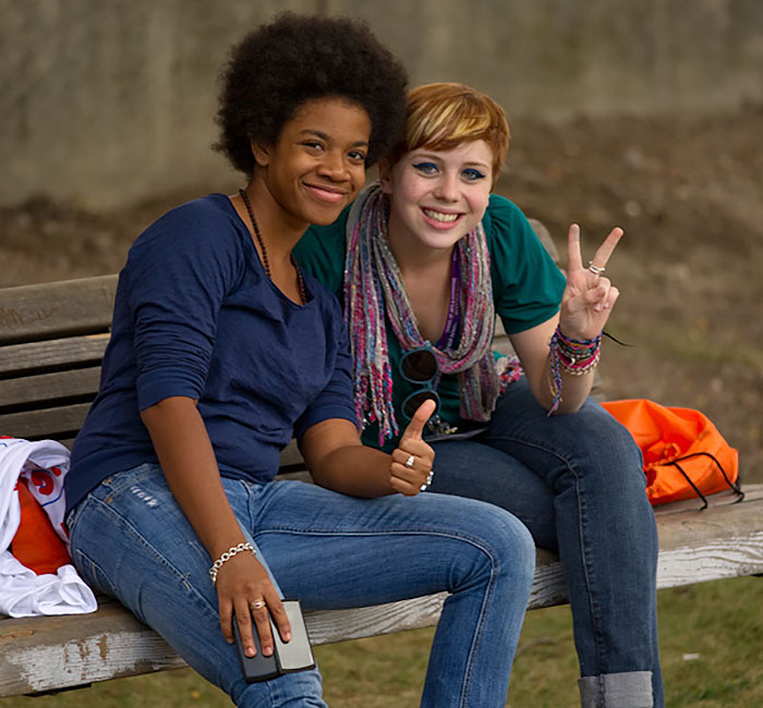 2 female college students sit on a bench outside smiling while one makes peace sign with her hand.