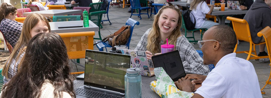 Binghamton University students sit at a cafe table outside with their laptops and talk.