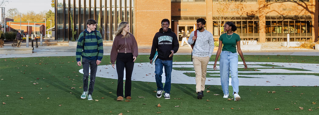 Five students talk while walking through a grass field in the middle of the Farmingdale State College campus.