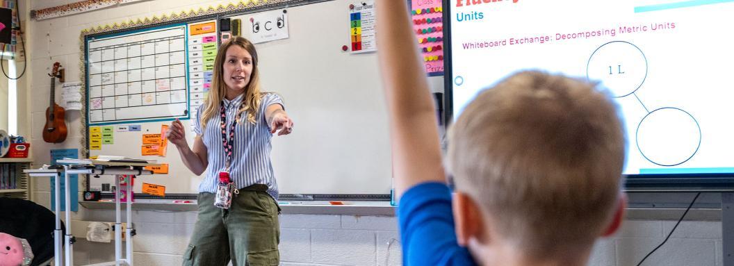 A female teacher points to a young boy while giving instruction in her elementary school classroom. 