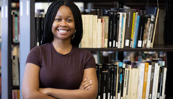 African American female with arms crossed stands in front of a book shelf in a library.