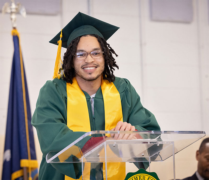 A student in green graduation cap and gown speaks at a glass podium.
