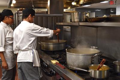 Two students of the SUNY Cobleskill culinary arts program stand over a chef's oven cooking a lunch meal in the school kitchen.	