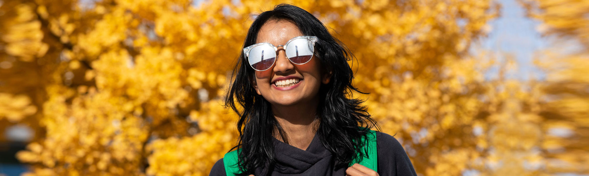 A female college student has a bright smile while wearing sunglasses and a black hoodie on a bright sunny day in front of a tree with yellow colored leaves.
