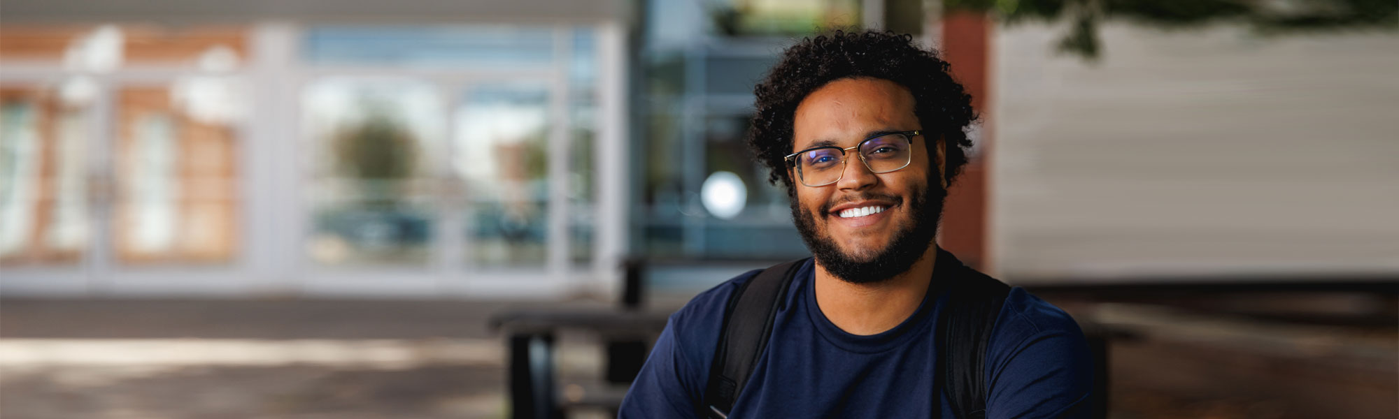 African american male student in dark blue tshirt sits on picnic table outside of college student center.