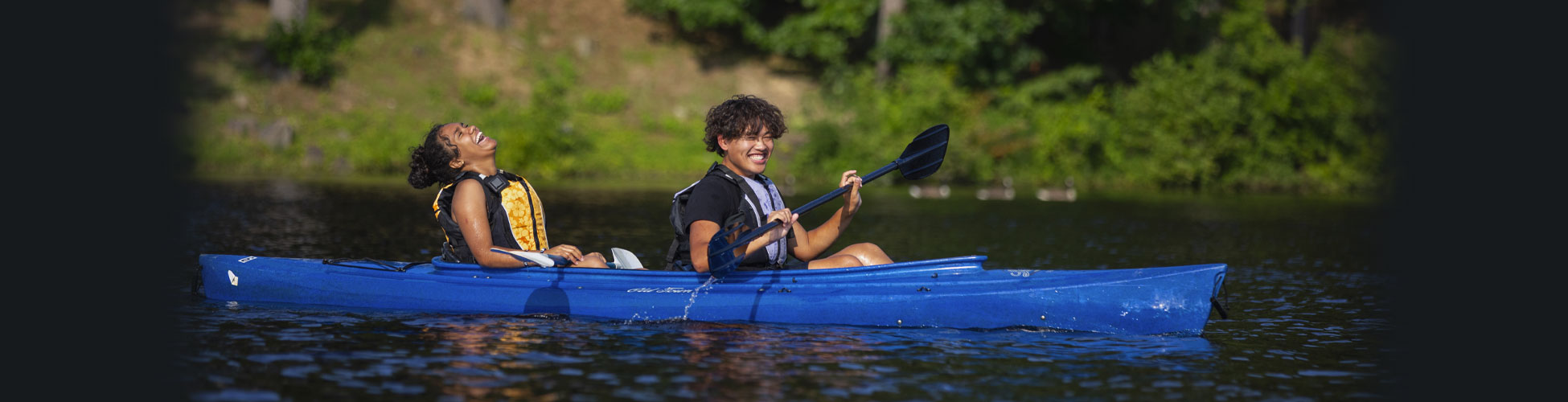 A male and female student laugh wildly while sitting in a canoe on a lake in updstate New York holding paddles up in their hands.