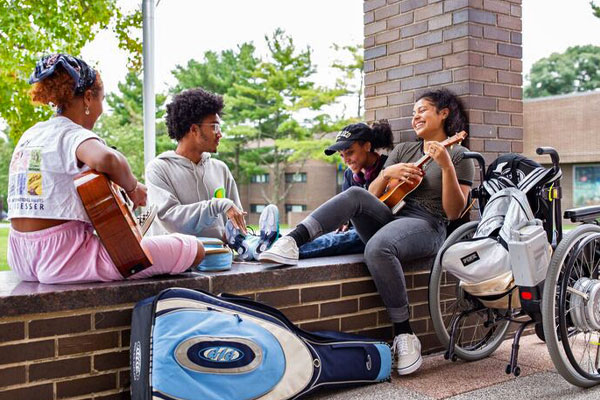 Diverse group of SUNY Purchase students sit on a brick wall outside while one plays guitar.