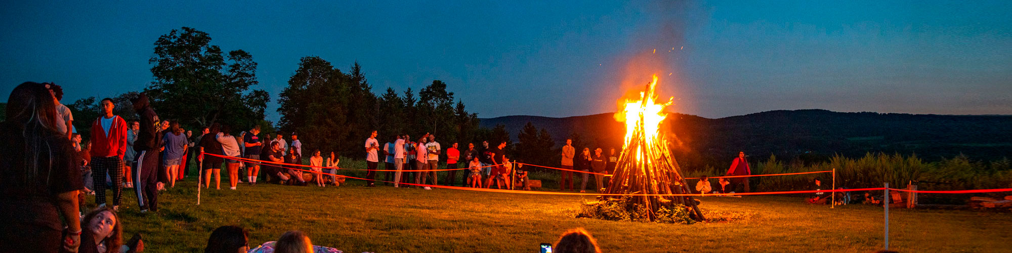 A bonfire in a large field surrounded by college students.