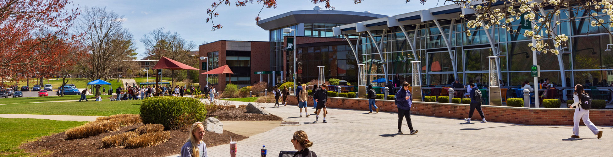 Students sit at table talking and other walk to and from the building on a bright fall day on the Binghamton University campus quad.
