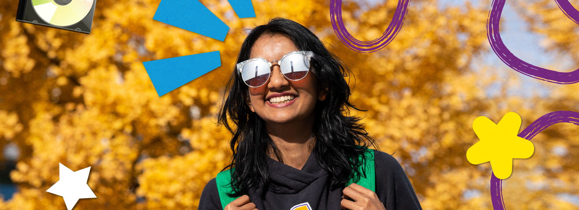 A female college student has a bright smile while wearing sunglasses and a black hoodie on a bright sunny day in front of a tree with yellow colored leaves.