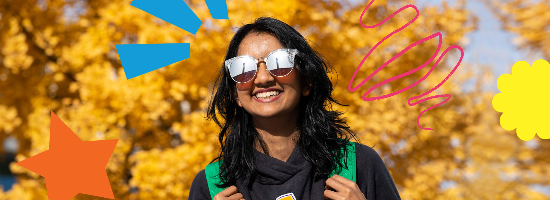 A female college student has a bright smile while wearing sunglasses and a black hoodie on a bright sunny day in front of a tree with yellow colored leaves.