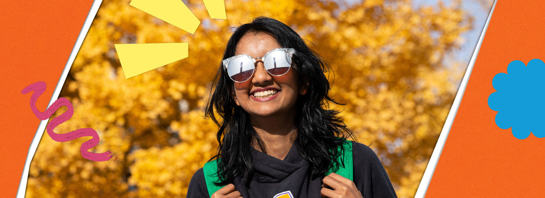 A female college student has a bright smile while wearing sunglasses and a black hoodie on a bright sunny day in front of a tree with yellow colored leaves.