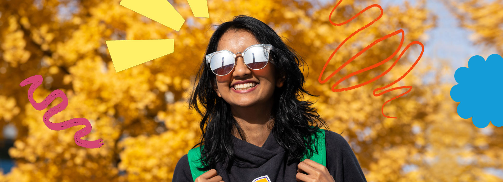 A female college student has a bright smile while wearing sunglasses and a black hoodie on a bright sunny day in front of a tree with yellow colored leaves.