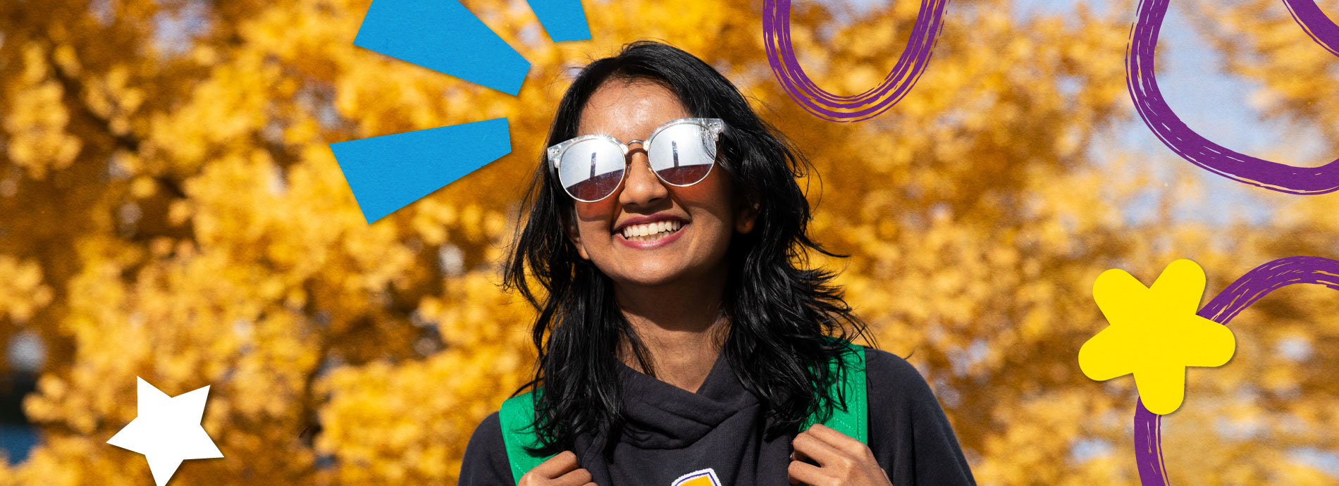 A female college student has a bright smile while wearing sunglasses and a black hoodie on a bright sunny day in front of a tree with yellow colored leaves.