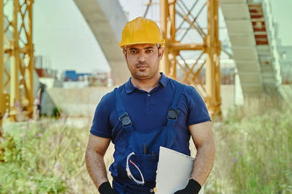 Female in hard hat and safety vest smiles outside in front of trees.