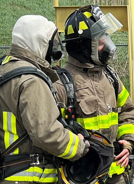 Two firefighters with full firefighter safety gear and helmets on look off to the side