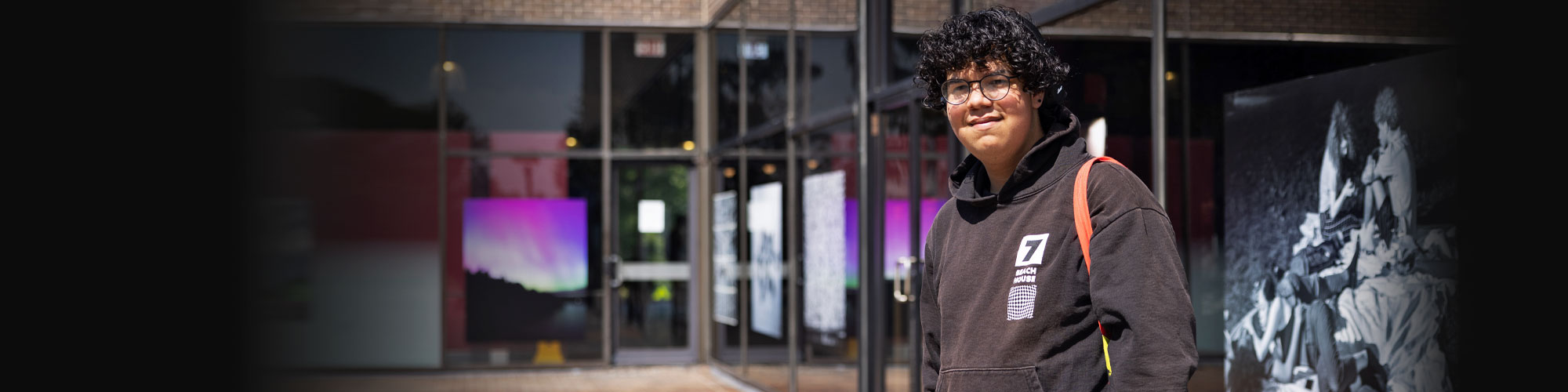 A male student with curly hair stands outside while wearing a black hoodie. 
