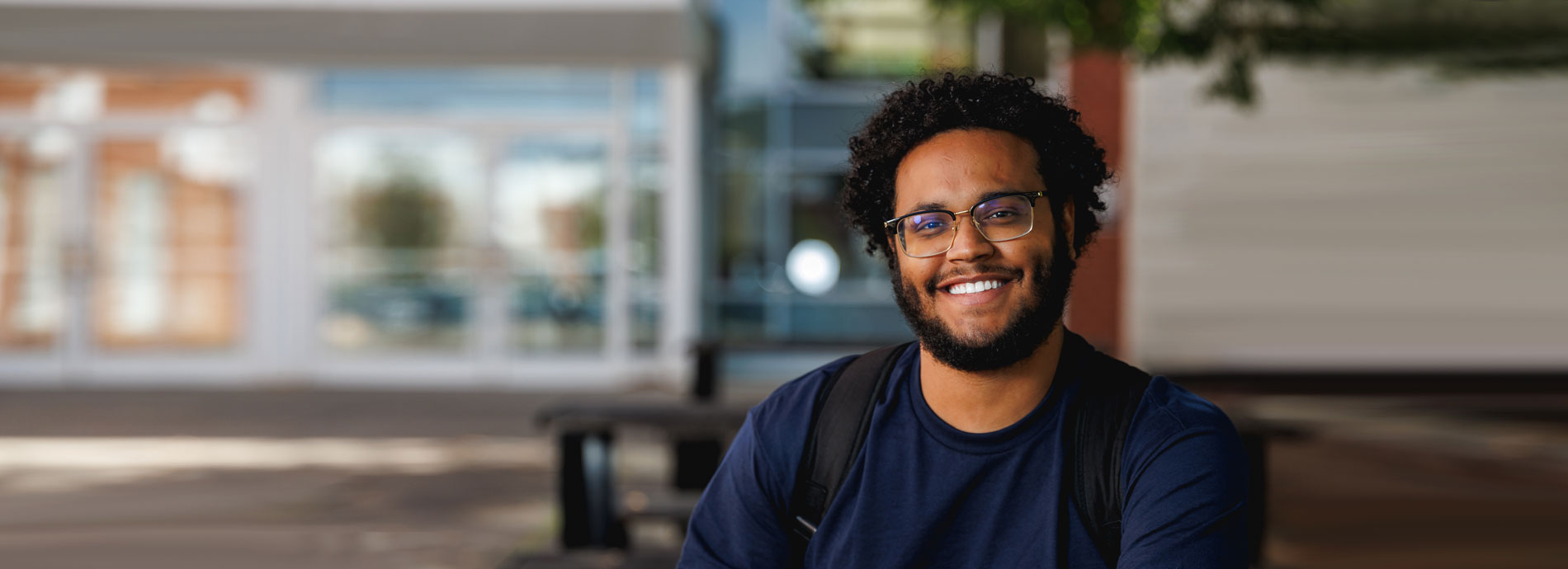 Afircan American male student in blue tshirt sits on picnic table outside in front of the SUNY Broome student center.