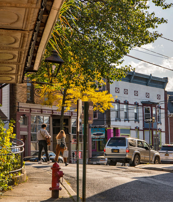 Tree leaves overhang a store awning as two people walk away to cross a small village street