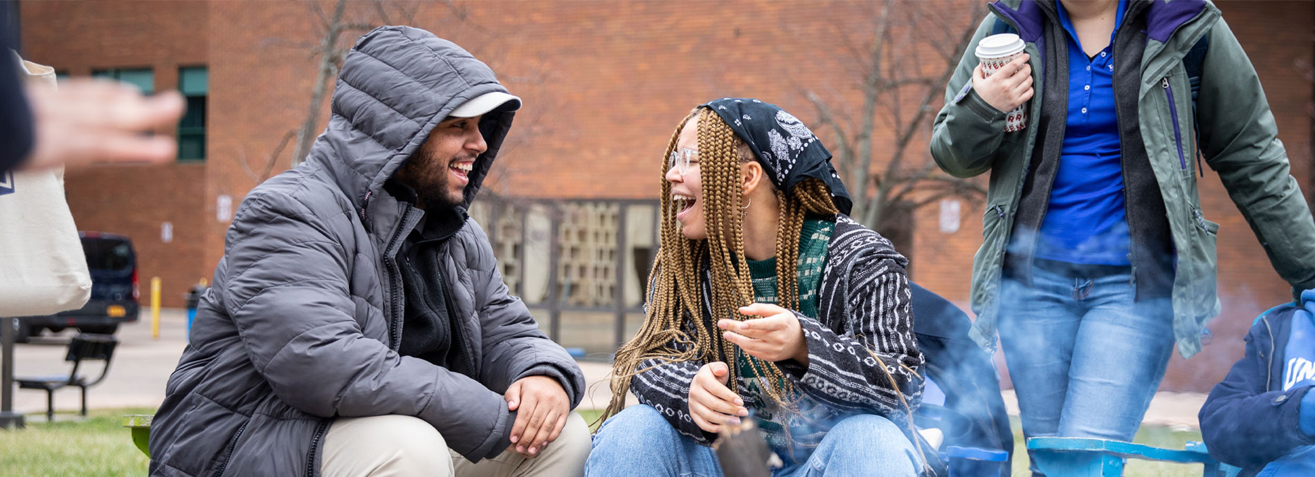 A male and female college student sit and talk behind a small fire pit.