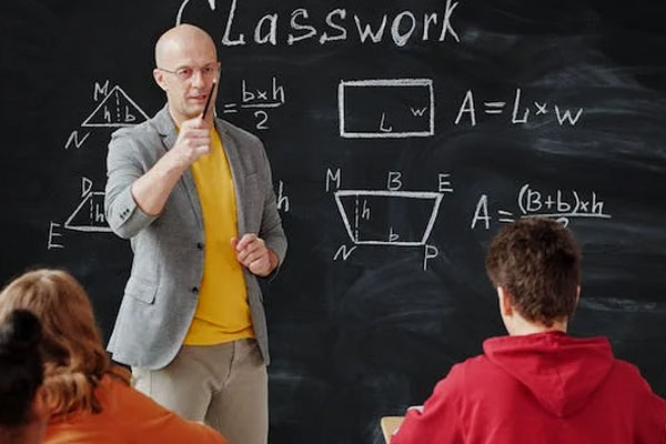 A male teacher standing in front of chalkboard with math equations on it points to a student.