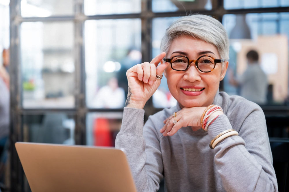 Asian woman with glasses works on a laptop in a common space.