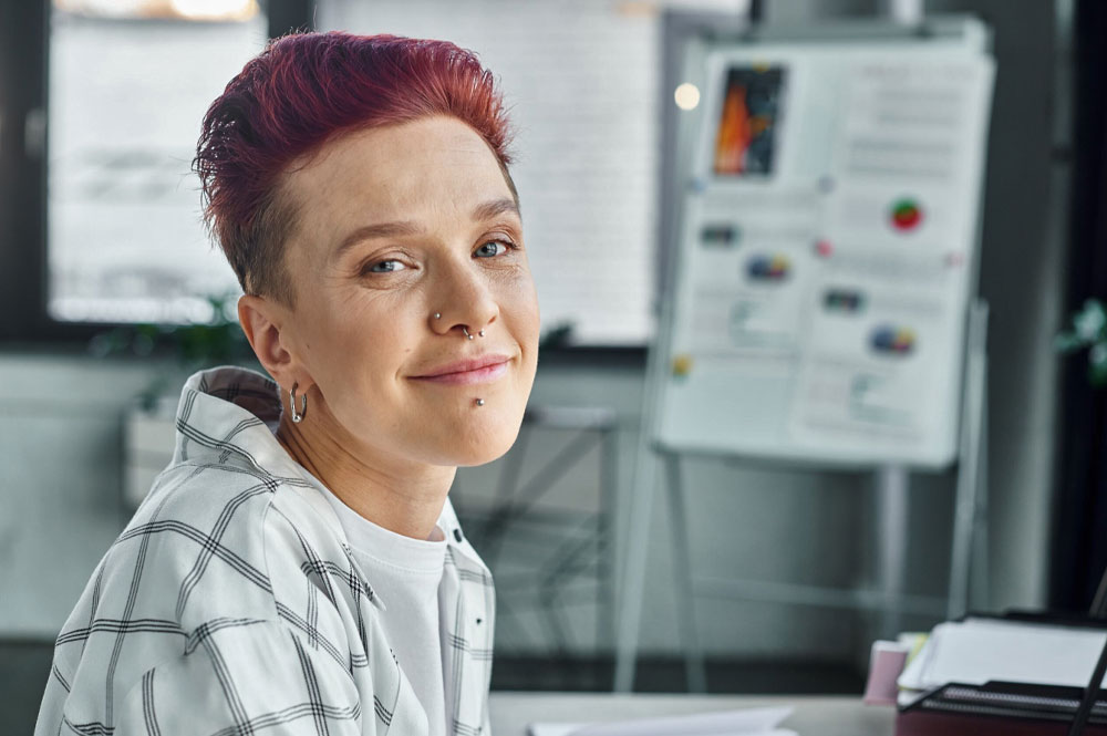 A female with short hair and nose ring sits in front of a business presentation on white board.
