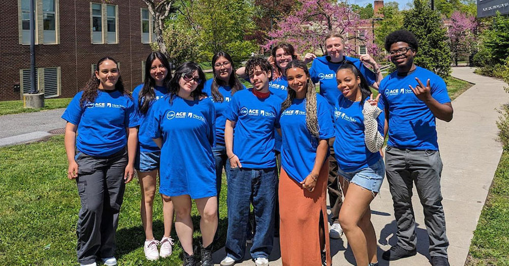Students all wearing blue SUNY ASAP ACE tshirts pose for a picture outside on the SUNY New Paltz campus.