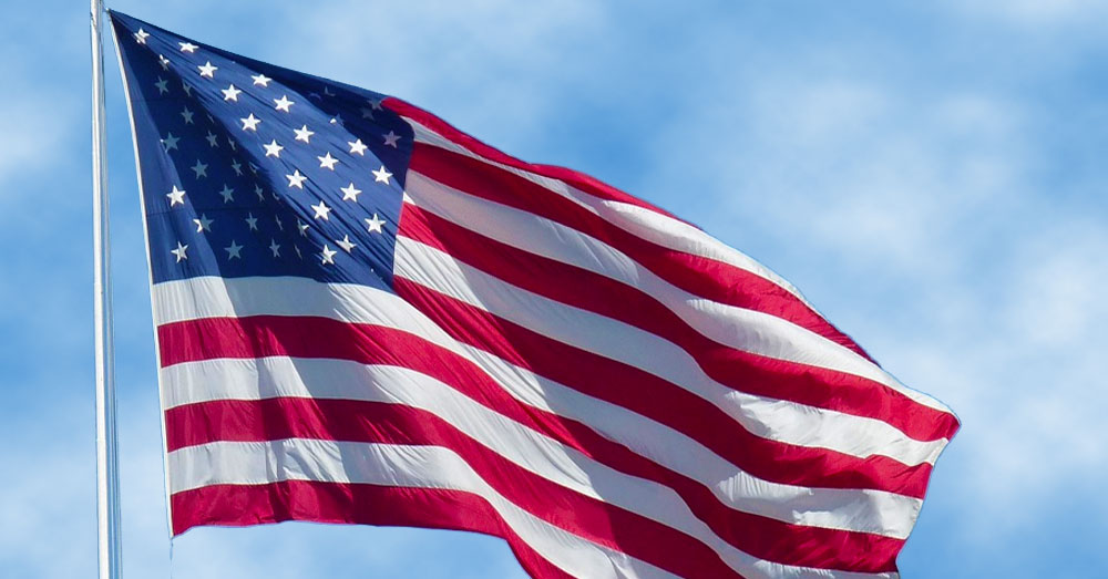 An American Flag flies with a bright cloudy sky behind it.