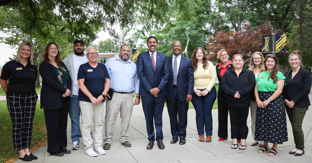 SUNY Chancellor John B. King Jr. stands with members of the SUNY Broome Community College community outside on a walkway under trees.