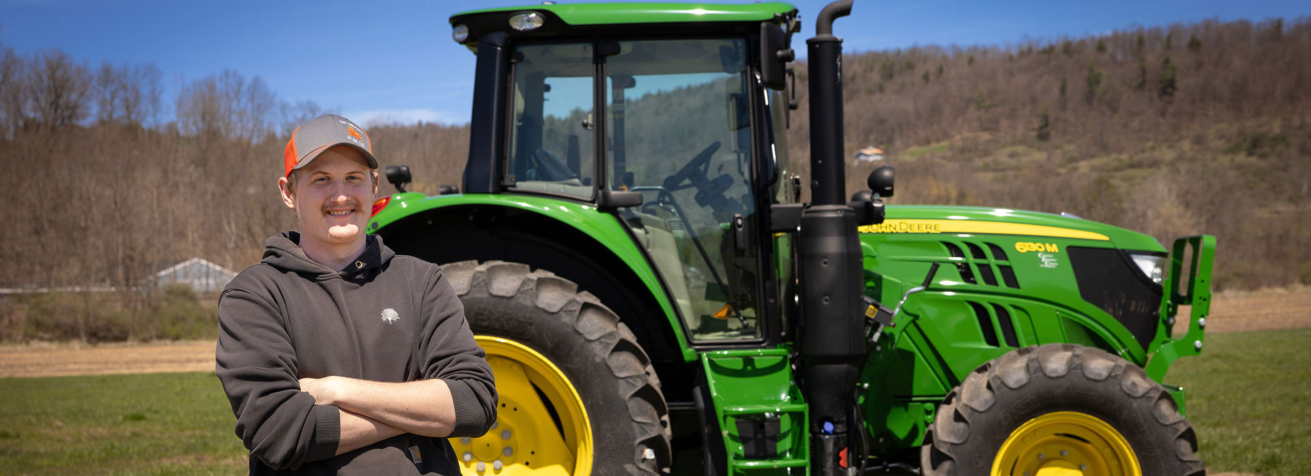 Male student wearing a gray hoodie stands in front of a large green John Deere tractor.