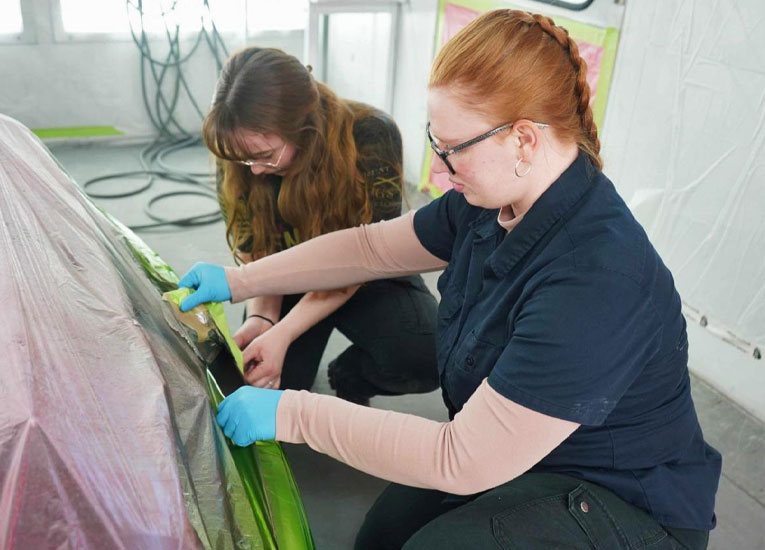 Two female students work on fixing a car body in a garage on the campus of SUNY Morrisville.