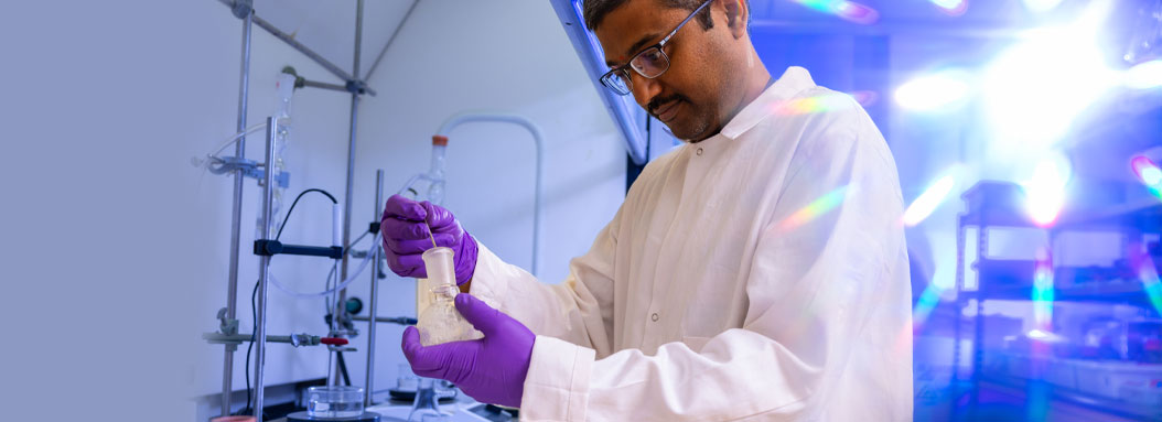A male research scientist holds a beaker in a lab while wearing a white lab coat.