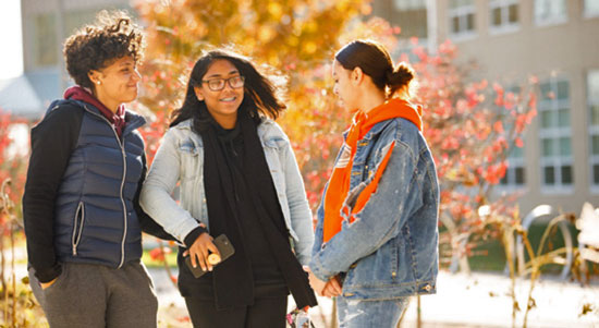 3 female college students talk outside during autumn on college campus.