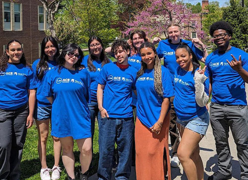 Students all wearing blue SUNY ASAP ACE tshirts pose for a picture outside on the SUNY New Paltz campus.