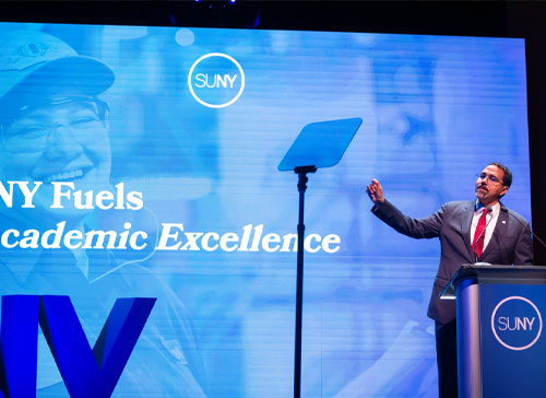 SUNY Chancellor John B. King Jr. delivers the 2026 State of the University Address with a screen behind him showing a slide with SUNY Fuels Academic Excellence written on it.