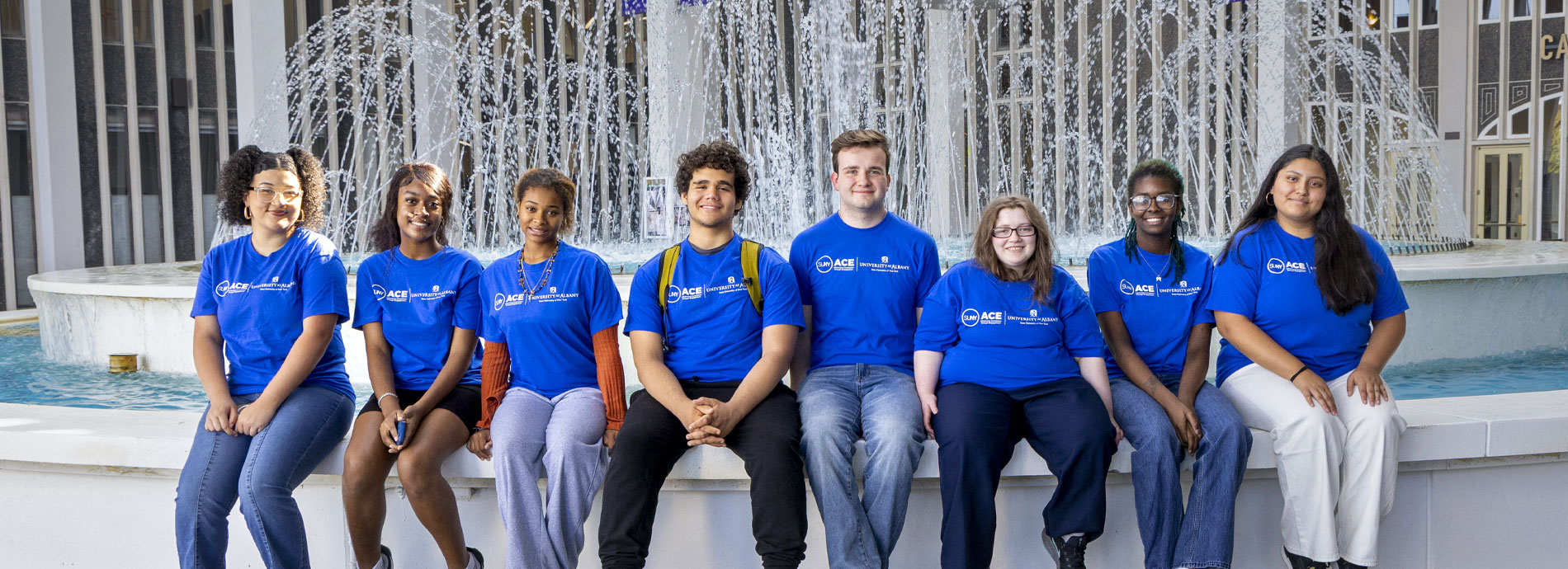 A group of UAlbany students sit on a ledge in front of the campus fountain all wearing blue SUNY ACE tshirts.