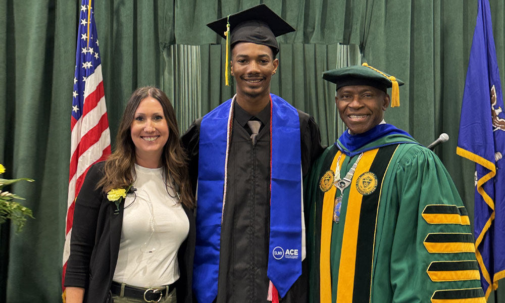 An african american male stands with SUNY Oswego president Peter Nwosu and another female for a picture in his college commencement robe and a SUNY ACE sash ribbon around his neck draped down his shoulders.