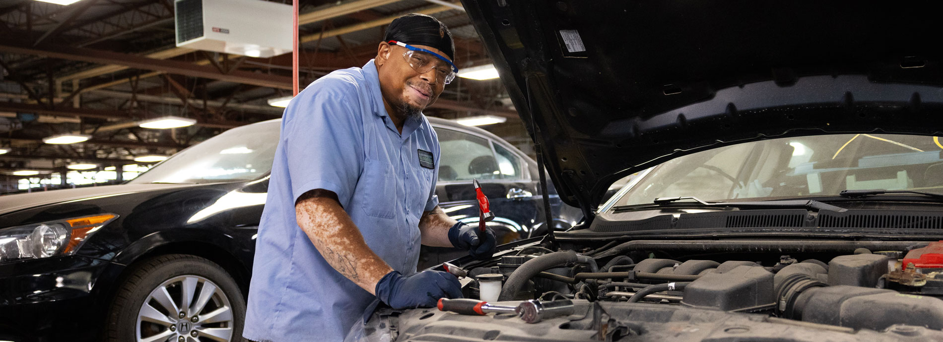 Man smiles while working under the hood of a car in a garage at Hudson Valley Community College.