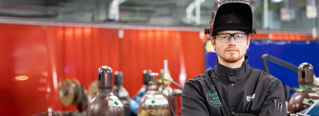 A male student in an auto repair training garage stands in front a group of air tanks while wearing a welding helmet.